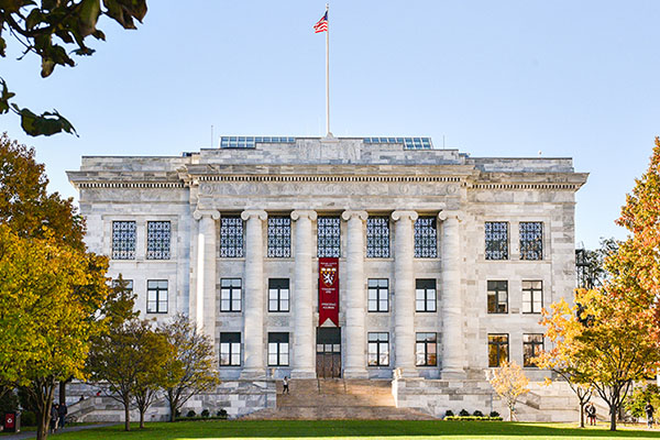 Colorful photograph of Gordon Hall on the HMS campus, a marble building with columns. In front is a manicured lawn and autumnal trees