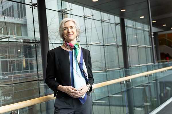 portrait photo of Jennifer Doudna, a slightly smiling white woman with chin-length gray hair, leaning against a railing in front of a wall of windows. she wears a blazer and colorful silk scarf