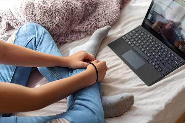Her face offscreen, a young woman sits on a couch with her arms and legs crossed, wearing ripped blue jeans. At her feet, a laptop shows a face, the details obscured by sun glare. A telemedicine therapy session is underway.